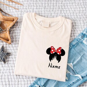 May include: Cream-colored t-shirt featuring a Minnie Mouse design with a red and white polka dot bow, black ears, and a castle silhouette. The word "Name" is written below the design. Light blue denim jeans are visible.
