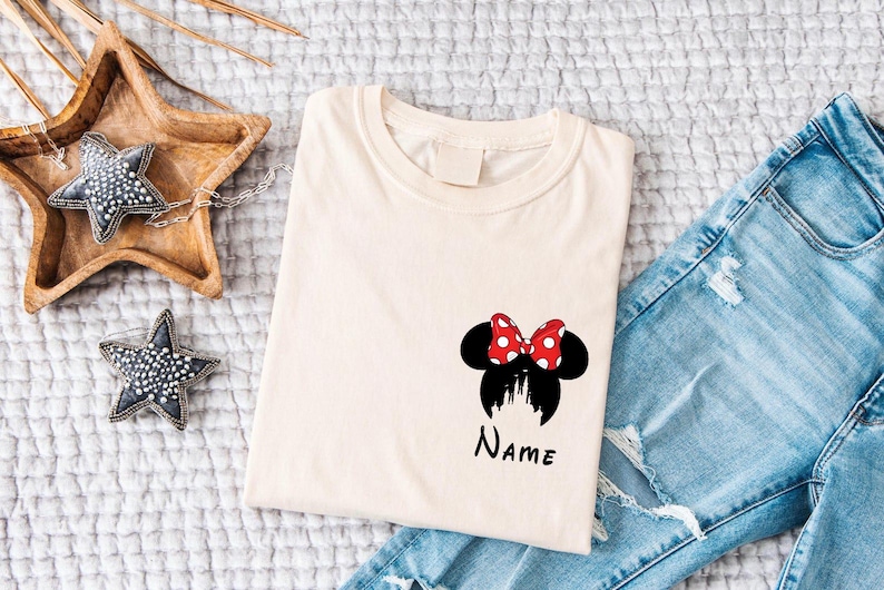 May include: Cream-colored t-shirt featuring a Minnie Mouse design with a red and white polka dot bow, black ears, and a castle silhouette. The word "Name" is written below the design. Light blue denim jeans are visible.
