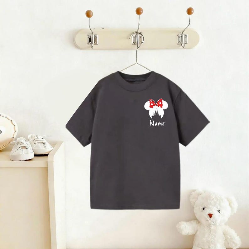 May include: Dark gray t-shirt with a white graphic of a bow and silhouette of a castle. The bow is red with white polka dots. The word "Name" is printed in white below the graphic. The shirt is hanging on a white wall.