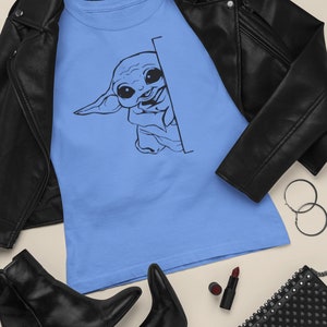 May include: A light blue t-shirt with a black and white illustration of a cartoon character peeking from behind a corner. The character has large eyes and a small mouth. The t-shirt is laid out on a light brown surface with a black leather jacket, black boots, a black purse, and a red lipstick.