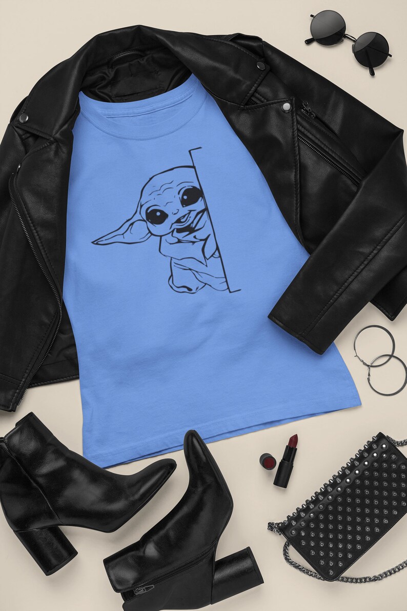 May include: A light blue t-shirt with a black and white illustration of a cartoon character peeking from behind a corner. The character has large eyes and a small mouth. The t-shirt is laid out on a light brown surface with a black leather jacket, black boots, a black purse, and a red lipstick.