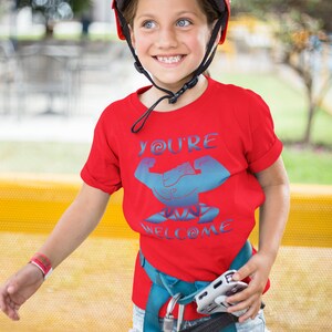 May include: A red t-shirt with a blue graphic of a bicep flexing with the text "You're Welcome" on it. The shirt is being worn by a child wearing a blue helmet.