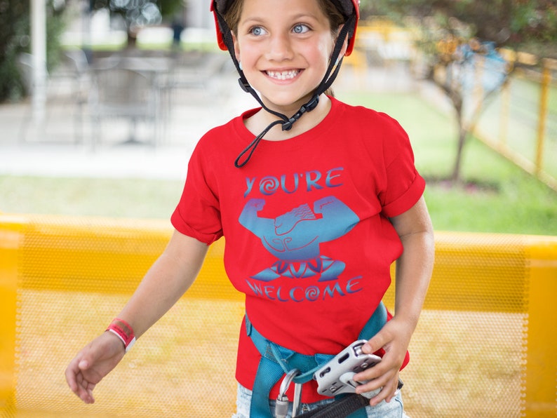 May include: A red t-shirt with a blue graphic of a bicep flexing with the text "You're Welcome" on it. The shirt is being worn by a child wearing a blue helmet.