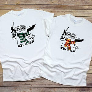 May include: Two white t-shirts with a graphic of a baby Yoda character wearing glasses and a scarf. One shirt has a green scarf and the other has a red and yellow scarf.