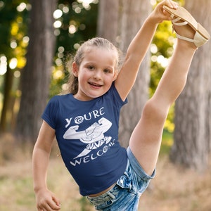 May include: A young person wearing a navy blue t-shirt with a white graphic of a cartoon character with the text "You're Welcome". The person is wearing denim shorts and has their leg raised in the air.