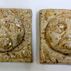 May include: Two ceramic tiles with a raised, textured design of a face. The tiles are a light beige color with brown and white accents.