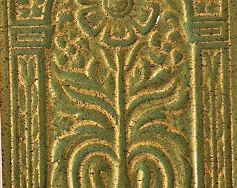May include: A green ceramic tile with a raised design of a flower and leaves in an archway. The tile has a speckled glaze.