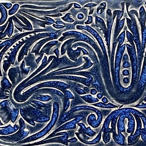 May include: Close-up of a decorative tile with a detailed floral pattern. The design features swirling leaves and stylized flowers in shades of blue and white. The tile's surface has a textured appearance, enhancing the intricate details.