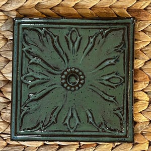 May include: Square, ceramic coaster with a floral design. The coaster is a mottled teal color with a dark brown border. The floral design features a central circle with smaller circles around it and stylized leaves.