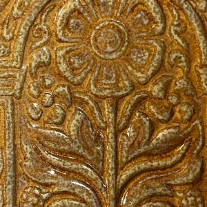May include: Close-up of a ceramic tile with a raised floral design. The tile features a central flower with detailed petals and a stem, surrounded by ornate, swirling patterns. The color palette includes shades of brown, beige, and gold, creating a textured, antique appearance.