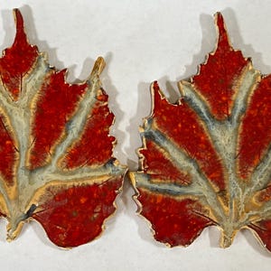May include: Two ceramic leaf-shaped ornaments with a red and white glaze. The leaves have a detailed vein pattern and a speckled texture.