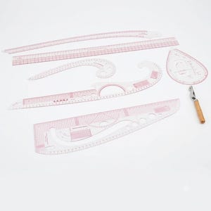 Può includere: Un set di righelli da cucito e strumenti per modellistica in plastica trasparente con segni rossi e misure in centimetri. La collezione include vari modelli curvi, un righello dritto, una guida a forma di goccia e un taglia asole in metallo.