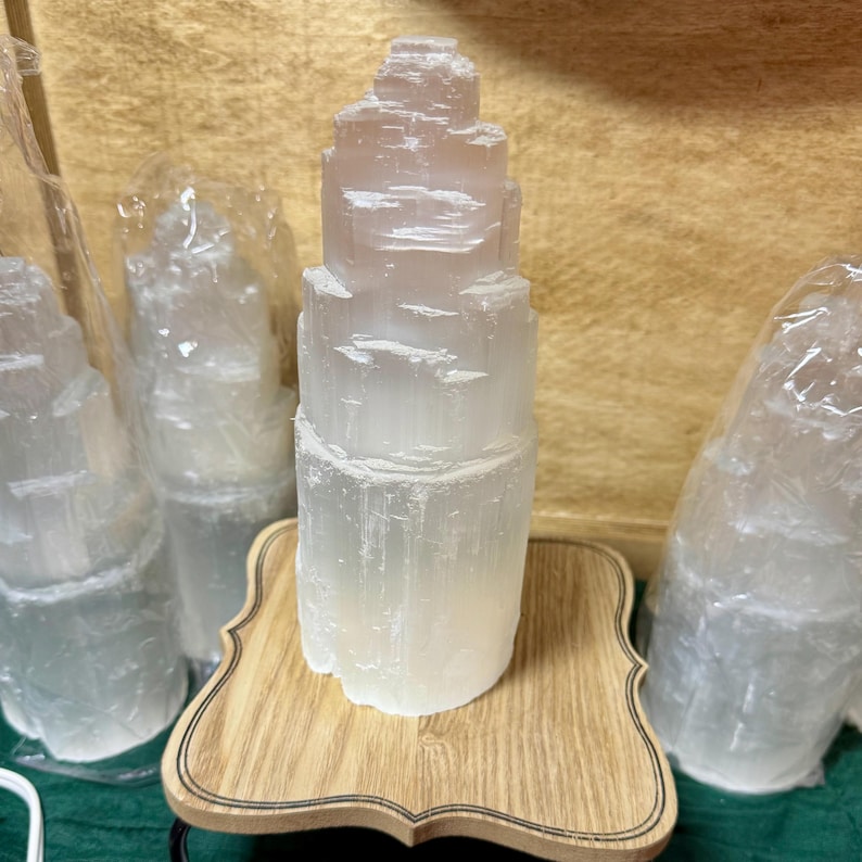 May include: A set of four white selenite crystal towers on a wooden base. The crystals are stacked in a tiered formation and have a natural, rough texture.