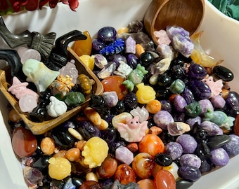 Pode incluir: Uma tigela branca cheia de uma variedade de pedras coloridas, cristais e pequenas estatuetas. As pedras são em tons de roxo, vermelho, verde e preto. As estatuetas têm a forma de morcegos, abóboras e outros objetos com tema de Halloween.