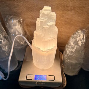 May include: A white selenite crystal tower lamp on a digital scale that reads 2.153. The scale is on a wooden surface. The lamp is lit and glowing.