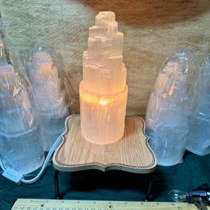 May include: A selenite crystal lamp with a wooden base and a warm glow. The lamp is made of several stacked selenite crystals and is lit from within. The lamp is surrounded by other selenite crystals.