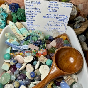 May include: A white bowl filled with a variety of colorful stones and crystals, including blue lace agate, trolleite, blue chalcedony, Caribbean calcite, aquamarine, and more. A wooden scoop is in the bowl, and a handwritten note lists the types of stones and crystals included. The note also says "Life's A Beach" and "confetti".