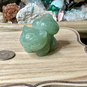 May include: A light green crystal figurine of a cartoon character, with a large head and bulbous back. The figurine is on a wooden surface, with a quarter coin to the left. The background features various crystals and minerals.