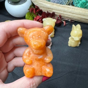 May include: A carved orange bear figurine is held in a hand, with other small animal figurines in the background. The bear is detailed with rounded features and a smooth surface. The figurines are likely made of stone or a similar material.
