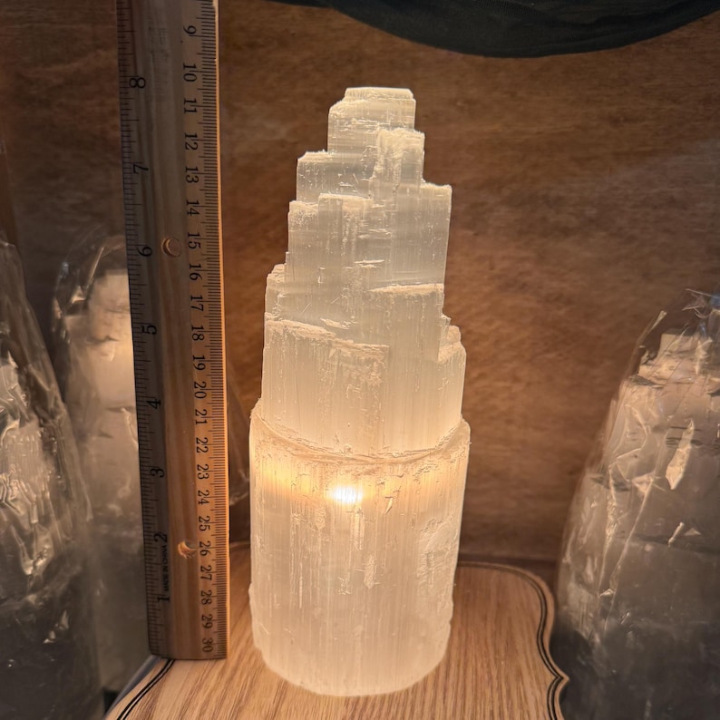 May include: A white selenite crystal lamp with a stacked, layered design. The lamp is lit and glowing, casting a warm light. The lamp is sitting on a wooden base.