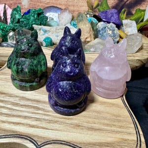 May include: Three small, hand-carved stone figurines of a cartoon character. The figurines are made of green, purple, and pink stone.