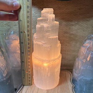 May include: A white selenite crystal lamp with a stacked tower design. The lamp is lit and glowing, casting a warm light. The lamp is approximately 18 cm tall.