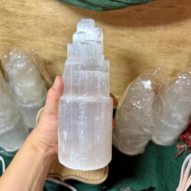 May include: A white selenite crystal tower with a layered, stacked design. The crystal is smooth and translucent, with a slightly cloudy appearance. The tower is sitting on a wooden base.