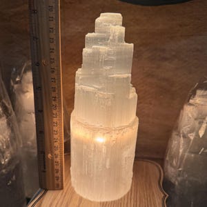 May include: A white selenite crystal tower with a warm glow emanating from the base. The tower is approximately 15 cm tall and is made of stacked, rectangular crystals.