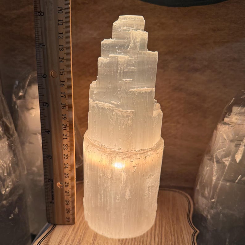 May include: A white selenite crystal tower with a warm glow emanating from the base. The tower is approximately 15 cm tall and is made of stacked, rectangular crystals.