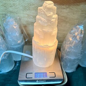May include: A white selenite crystal lamp with a stacked tower design. The lamp is lit and casting a warm glow. The lamp is on a digital scale that reads 370.1.
