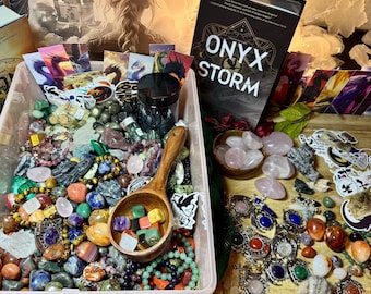 May include: A pink plastic container filled with a variety of colourful stones, crystals, and beads. There are also several bracelets and necklaces in the container. The container is sitting on a wooden table next to a book titled "Onyx Storm".