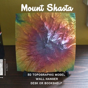 May include: A 3D topographic model of Mount Shasta, a volcano in California. The model is made of a colorful material and is 6 inches in size. It is displayed on a black stand and can be used as a wall hanger or desk decoration.