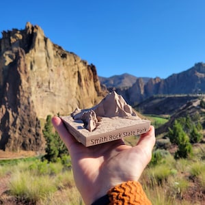 May include: A 3D printed model of Smith Rock State Park in Oregon, held in a hand against a backdrop of the actual rock formations. The model is made of a light brown material and shows the distinctive rock formations of the park.