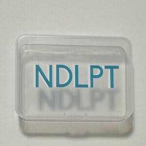 May include: A clear, rectangular plastic container with a hinged lid. The container has the teal letters "NDLPT" printed on the front. The letters are also faintly visible as a shadow beneath the primary text.