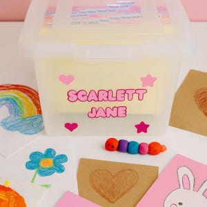May include: A clear plastic storage bin with a lid, labeled "SCARLETT JANE" in pink letters. The bin is decorated with pink hearts and stars. Artwork, including drawings of a rainbow, flowers, and a bunny, surrounds the bin.