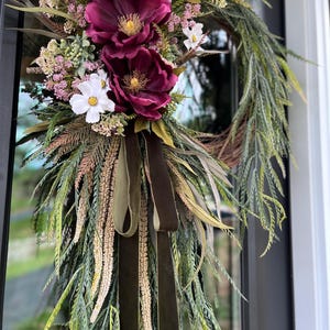 May include: A teardrop-shaped floral wreath featuring deep burgundy peonies, white cosmos flowers, and small pink filler blossoms. The arrangement includes long, trailing green foliage, wispy grasses, and a dark olive green velvet ribbon bow.