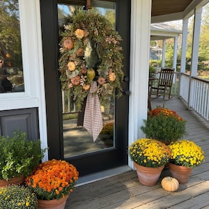 May include: A decorative wreath featuring green foliage, small orange and yellow pumpkins, and a hanging brown and white checkered fabric ribbon, suspended by a brown leather strap on a black glass-paneled door with a wooden porch setting.