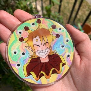 Könnte beinhalten: Ein runder, klarer Acryl-Charm mit einem lächelnden Cartoon-Charakter. Der Charakter hat gelbes Haar, einen braunen Rüschenkragen und einen Hintergrund in Pastellfarben. Oben befindet sich eine kleine lila Schlaufe.