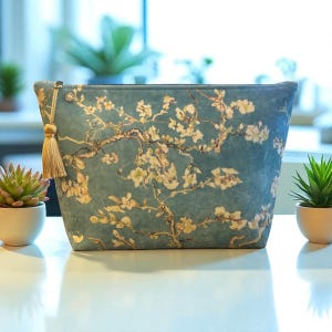 May include: A teal cosmetic bag featuring a floral design reminiscent of Van Gogh's "Almond Blossoms." The bag has a gold tassel zipper pull. The bag is sitting on a white surface.