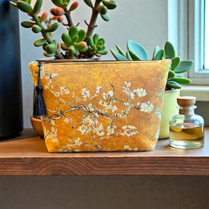 Puede incluir: Un neceser amarillo con un diseño floral inspirado en "Almendros en flor" de Van Gogh. La bolsa tiene una cremallera y una borla negras. La bolsa está sobre una estantería de madera con plantas y una pequeña botella de vidrio.