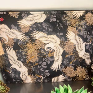 May include: Black zippered pouch with a gold tassel. The pouch features a repeating pattern of white cranes and gold floral designs on a black background.