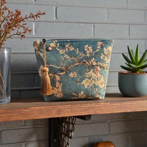 May include: Teal velvet cosmetic bag with a floral design inspired by Van Gogh's Almond Blossoms. It has a gold tassel zip pull and is on a wooden shelf. A small succulent in a blue pot is also visible.