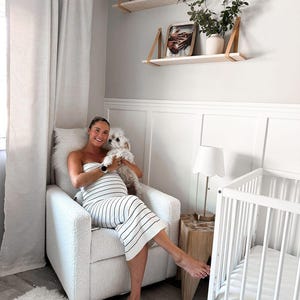 Puede incluir: Una mujer embarazada se sienta en un sill&oacute;n blanco con un peque&ntilde;o perro blanco en sus brazos. La mujer lleva un vestido de rayas blancas y negras. Hay una cuna blanca con un colch&oacute;n blanco en el fondo. La habitaci&oacute;n est&aacute; decorada en un estilo minimalista con paredes blancas y estantes de madera.
