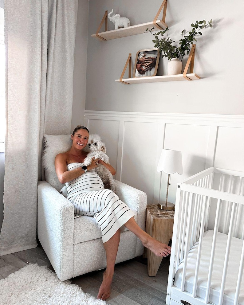 Puede incluir: Una mujer embarazada se sienta en un sill&oacute;n blanco con un peque&ntilde;o perro blanco en sus brazos. La mujer lleva un vestido de rayas blancas y negras. Hay una cuna blanca con un colch&oacute;n blanco en el fondo. La habitaci&oacute;n est&aacute; decorada en un estilo minimalista con paredes blancas y estantes de madera.