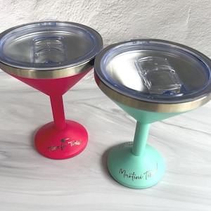 May include: Two stainless steel martini glasses with clear plastic lids. One glass is hot pink with the text Time for Tinis! and the other is mint green with the text Martini Time! printed in gold on the base of each stem.