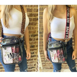 May include: Two clear plastic crossbody bags with different straps. One bag has a leopard print strap and the other has a red and white strap with the text "OU LOVE SEE YOU LOVE". Both bags have black trim and a zipper closure. The bag on the left has the initials "VME" in black letters on the front. The bag on the right has the initial "M" in red letters on the front.