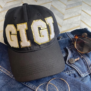 May include: Black baseball cap with a white felt patch that says "GIGI" in gold glitter letters.