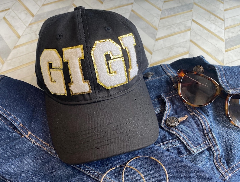 May include: Black baseball cap with a white felt patch that says "GIGI" in gold glitter letters.