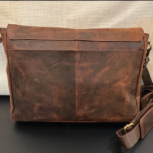 Pu&ograve; includere: Una borsa a tracolla in pelle marrone con patta rettangolare, una cucitura verticale al centro e una tracolla regolabile con una sezione imbottita. La borsa presenta finiture in metallo color ottone e una superficie testurizzata.