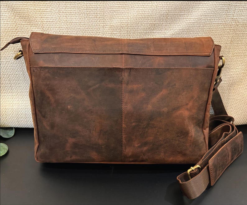 Pu&ograve; includere: Una borsa a tracolla in pelle marrone con patta rettangolare, una cucitura verticale al centro e una tracolla regolabile con una sezione imbottita. La borsa presenta finiture in metallo color ottone e una superficie testurizzata.